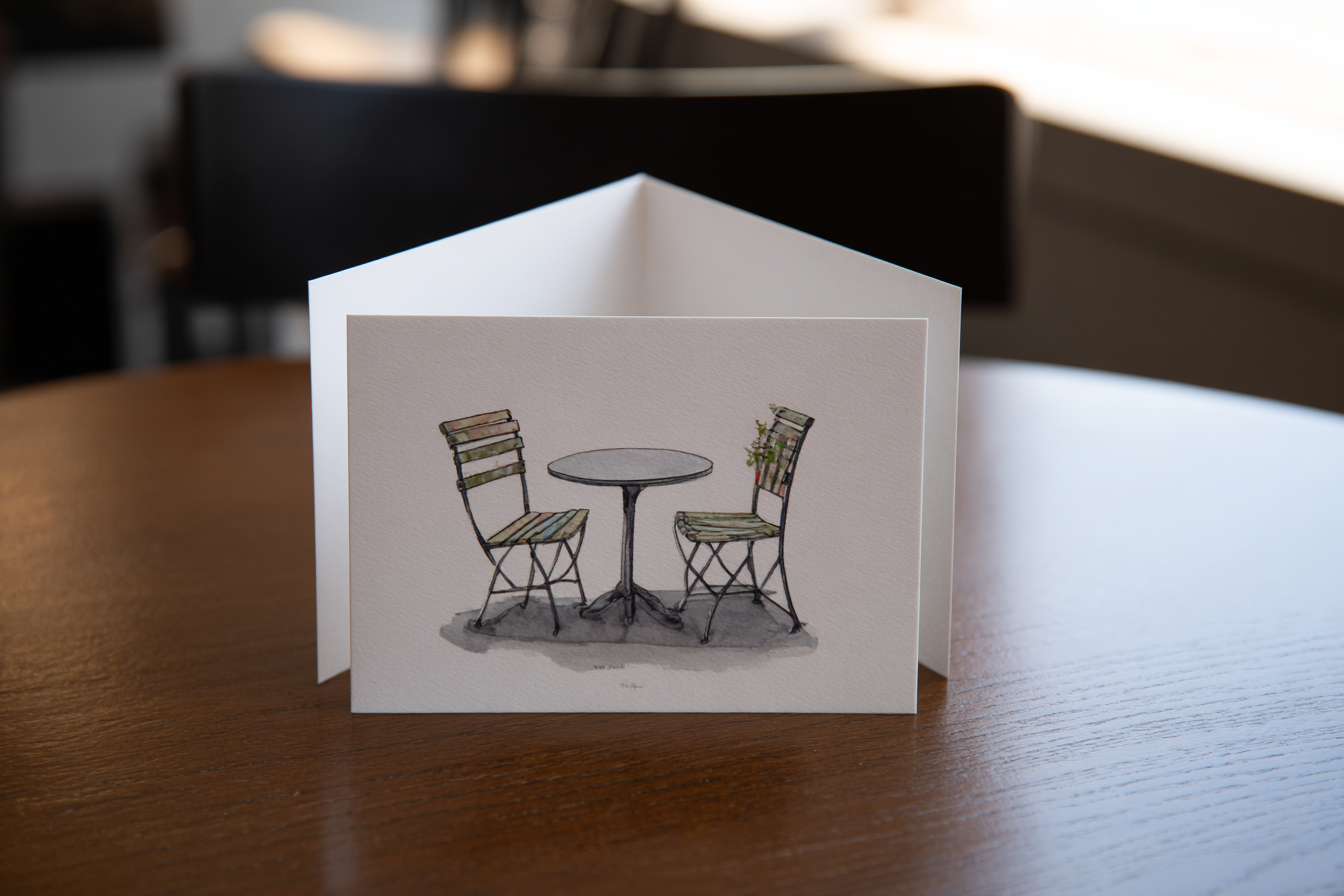 A greeting card lying flat, card front showing two empty chairs at a cafe table